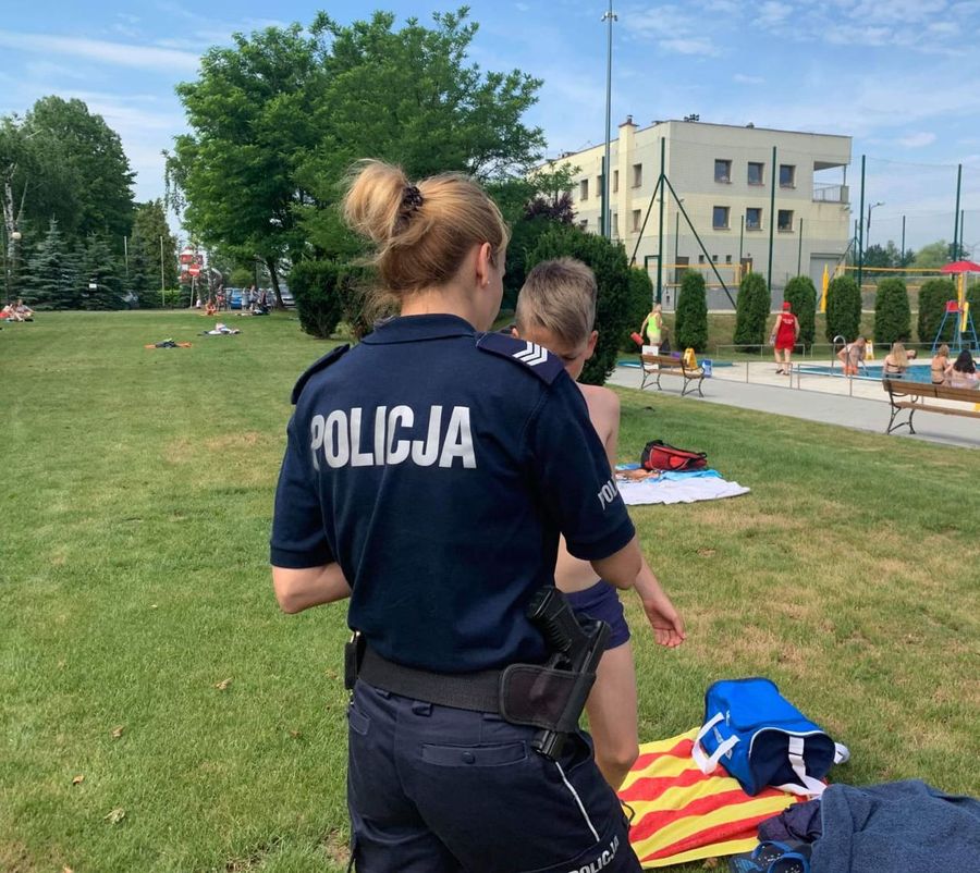 Fotografia kolorowa. Na zdjęciu widoczna policjantka stojąca tyłem wręczająca ulotkę dziecku.