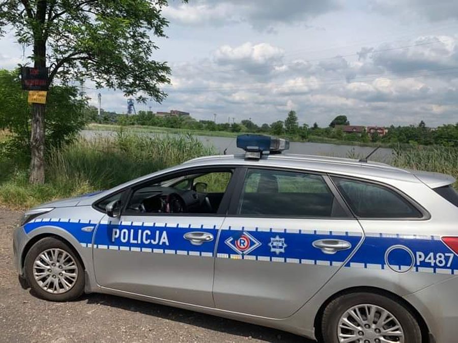 Fotografia kolorowa. Na zdjęciu widoczna policjantka stojąca obok radiowozu. W tle widoczny zbiornik wodny. 