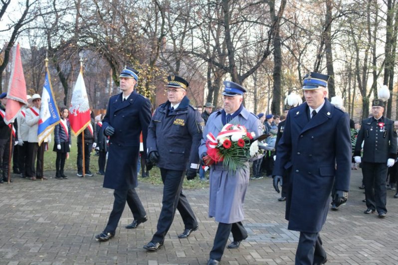 Fotografia kolorowa. Na zdjęciu widoczny Komendant wraz z przedstawicielami zaprzyjaźnionych służb. Delegacja zmierza przez pomnik aby złożyć przed nim kwiaty.