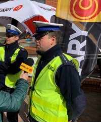 Zdjęcie kolorowe. Na zdjęciu widoczny policjant udzielający wywiadu dla radia piekary. Funkcjonariusz w odblaskowej kamizelce z napisem policja.