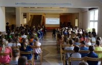 Fotografia kolorowa. Zdjęcie w trakcie zajęć przeprowadzanych w szkole. Widoczne są dzieci oraz goście z Kuby, Chille, Niemiec wraz z policjantką prowadzącą zajęcia. Na ekranie wyświetlane są poszczególne slajdy prezentacji.