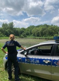 Fotografia kolorowa. Na zdjęciu widoczna policjantka stojąca obok radiowozu. W tle widoczny zbiornik wodny.