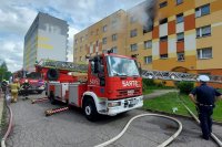 Miejsce pożaru mieszkania w budynku wielorodzinnym. Z lewej strony stoją dwa oznakowane pojazdy oraz funkcjonariusze straży pożarnej. Z prawej stoi umundurowany policjant.