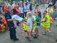 Policjantka stojąca obok radiowozu i rozdająca dzieciom kolorowanki- &quot;Sznupek kontra obcy&quot;.