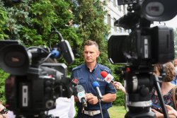 Zdjęcie kolorowe. Briefing prasowy przed komendą wojewódzką policji, przed mikrofonami głos zabiera umundurowany policjant.