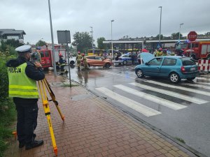 Umundurowany policjant podczas obsługi tachimetru na miejscu zdarzenia drogowego.  W tle pojazdy osobowe uczestniczące w zdarzeniu oraz pojazdy Policji, Straży Pożarnej oraz Pogotowia Ratunkowego