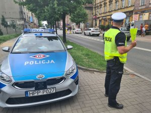 Umundurowany policjant ruchu drogowego stojący przy jezdni obok oznakowanego radiowozu i trzymający w ręku odblaskowe opaski.