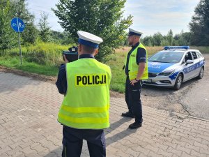 Dwaj umundurowani policjanci ruchu drogowego stojący na chodniku i kontrolujący prędkość pojazdów. W tle oznakowany policyjny radiowóz.