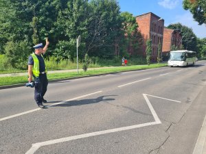 Umundurowany policjant wydziału ruchu drogowego stojący na środku jezdni i dający za pomocą dłoni sygnał do zatrzymania kierującemu autobusem miejskim.