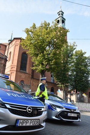 Umundurowany policjant ruchu drogowego stojący obok oznakowanych radiowozów na tle piekarskiej bazyliki