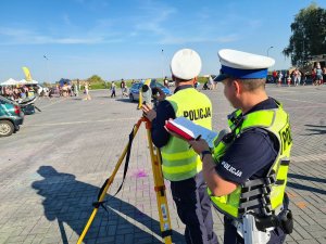 Umundurowani policjanci ruchu drogowego podczas pokazu zdarzenia drogowego. Jeden policjant wypełnia protokół oględzin pojazdu, natomiast drugi policjant obsługuje urządzenie tachimetr. W tle rozbite pojazdy oraz widownia.