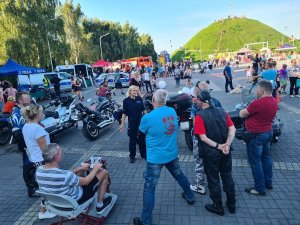 Policyjna profilaktyk podczas prelekcji z motocyklistami będącymi na pikniku. W oddali zaparkowane motocykle oraz Kopiec Wyzwolenia