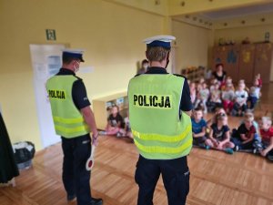 Umundurowani policjanci ruchu drogowego podczas spotkania z uczniami. Z tyłu Po lewej stronie grupa dzieci siedząca na podłodze. Po prawej  grupa dzieci siedząca na podłodze.