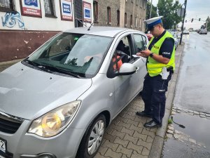 Umundurowany policjant ruchu drogowego stojący obok kontrolowanego pojazdu. Policjant trzyma w ręku tarczę do zatrzymywania pojazdów oraz ulotki promujące Europejski Tydzień Mobilności. Z tyłu policyjny radiowóz