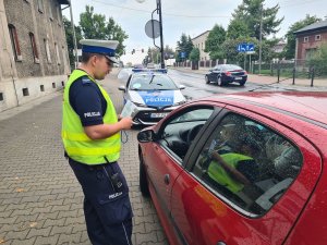 Umundurowany policjant ruchu drogowego stojący obok kontrolowanego pojazdu. Policjant trzyma w ręku tarczę do zatrzymywania pojazdów oraz ulotki promujące Europejski Tydzień Mobilności. Z tyłu policyjny radiowóz