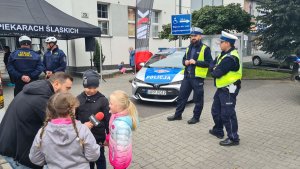 Po lewej mężczyzna rozmawiający z dziećmi, z tyłu przedstawiciele Straży Miejskiej, po prawej stronie umundurowani policjanci stojący obok radiowozu