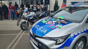 Policyjny motocykl oraz radiowóz na którym poukładane są laurki wykonane przez przedszkolaków. W oddali uczestnicy spotkania profilaktycznego.