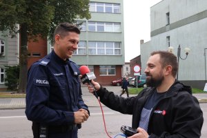 Po lewej stronie umundurowany policjant podczas wywiadu. Po prawej stronie mężczyzna trzymający mikrofon w ręce