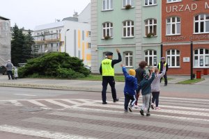 Umundurowany policjant oraz dzieci przechodzący przez przejście oraz trzymający podniesioną rękę. W oddali stoi drugi umundurowany policjant na tle budynku