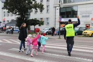 Umundurowany policjant przechodzący wraz z dziećmi przez przejście dla pieszych. Przed przejściem dla pieszych stoją samochody