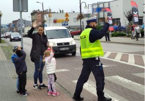 Umundurowany policjant przechodzący wraz z dziećmi przez przejście dla pieszych. Przed przejściem dla pieszych stoją samochody