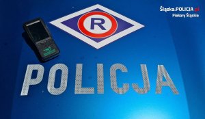 Zdjęcie pokrywy silnika policyjnego radiowozu, na której naklejony jest napis &quot;Policja&quot; oraz logo ruchu drogowego. Z boku położony jest narkotester.