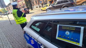 Umundurowany policjant ruchu drogowego stojący na chodniku obok oznakowanego radiowozu. Na bocznej szybie radiowozu naklejony plakat informujący o &quot;Ogólnopolskim Dniu Odblasków&quot;