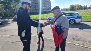 Umundurowany policjant ruchu drogowego stojący na chodniku wraz z mężczyzną oraz chłopczykiem. Policjant wręcza mężczyźnie opaskę odblaskową. W tle oznakowany policyjny radiowóz.