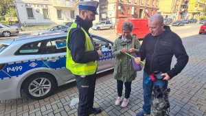 Umundurowany policjant ruchu drogowego stojący na chodniku wraz z kobietą i mężczyzną obok oznakowanego radiowozu. Na bocznej szybie radiowozu naklejony plakat informujący o &quot;Ogólnopolskim Dniu Odblasków&quot;. Policjant wręcza pieszym opaski odblaskowe.