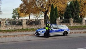 Umundurowany policjant stoi obok oznakowanego radiowozu. W tle cmentarz