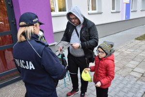 Umundurowana policjantka wręczająca mężczyźnie z dzieckiem ulotkę informacyjną oraz czapkę z daszkiem w jaskrawym kolorze promującą prowadzoną przez Śląską Policję kampanię &quot;Mamo! Tato! tutaj jestem&quot;