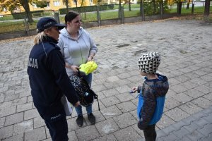 Umundurowana policjantka wręczająca kobiecie z dzieckiem ulotkę informacyjną oraz czapkę z daszkiem w jaskrawym kolorze promującą prowadzoną przez Śląską Policję kampanię &quot;Mamo! Tato! tutaj jestem&quot;