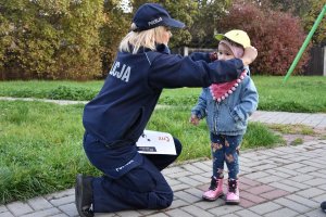 Umundurowana policjantka klęcząca przed dzieckiem i zakładająca na głowę dziewczynki czapkę z daszkiem w ramach prowadzonej przez Śląską Policję kampanii &quot;Mamo! Tato! tutaj jestem&quot;