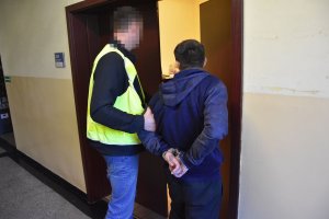 Policjant ubrany w odblaskową kamizelkę wprowadza zatrzymanego do pokoju.