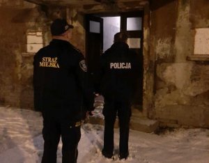 Umundurowany policjant wspólnie ze strażnikiem miejskim znajdują się przed budynkiem-kontrolują miejsca przebywania osób bezdomnych