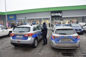 Umundurowany policjant oraz strażnik miejski stoją przy pojazdach służbowych na terenie centrum handlowego