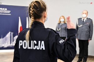 Nowo przyjęta policjantka podczas ślubowania trzyma w górze dwa palce, powtarzając słowa roty. Zdjęcie wykonane z tyłu, widoczny warkocz kobiety.