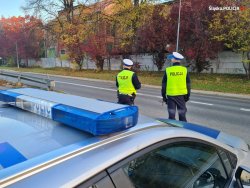 Na pierwszym planie policyjny radiowóz. W tle policjanci ruchu drogowego.