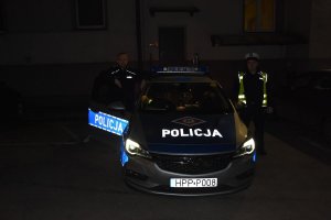 Dwaj umundurowani policjanci stoją przy oznakowanym radiowozie na parkingu komendy.