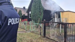 Zbliżenie na napis &quot;policja&quot; znajdujący się na kurtce policjantki. W tle dom jednorodzinny, w którym wybuchł pożar.