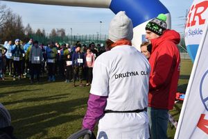 Kobieta ubrana w koszulkę z napisem &quot;Drużyna Olka&quot; stoi obok mężczyzny oraz policjantki. W tle uczestnicy biegu