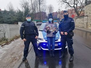 Dwóch umundurowanych policjantów wraz z wolontariuszką stoją przy oznakowanym radiowozie