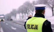 Umundurowany policjant w kamizelce odblaskowej z napisem POLICJA stoi przy krawędzi jezdni. W oddali porusza się samochód ciężarowy