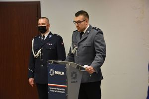 Umundurowany policjant w stroju galowym stoi przy mównicy. W tle Policjant w stroju galowym