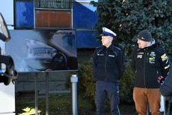 Policjant wraz z kierowcą rajdowym stoją na zewnątrz budynku. Obok nich p[ostawiony jest telewizor