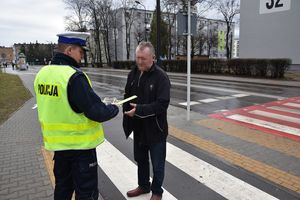 Umundurowany policjant w odblaskowej kamizelce wręcza mężczyźnie odblaskową opaskę. W tle jezdnia oraz przejście dla pieszych