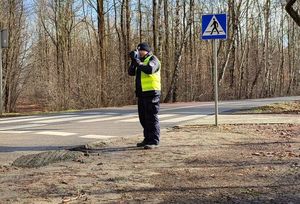 Umundurowany policjant w odblaskowej kamizelce stoi na poboczu przy oznakowanym przejściu dla pieszych i kontroluje prędkość pojazdów.