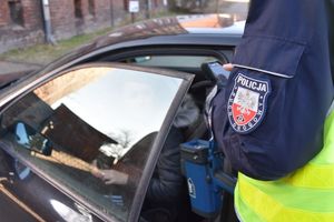 Umundurowany policjant w kamizelce odblaskowej stoi przy zatrzymanym do kontroli samochodzie. W ręce trzyma terminal mobilny oraz urządzenie do pomiaru prędkości