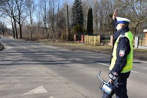 Umundurowany policjant w kamizelce odblaskowej zatrzymuje pojazd. W ręce trzyma urządzenie do pomiaru prędkości