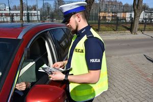 Umundurowany policjant stoi obok kontrolowanego pojazdu i wręcza kierującemu opaskę odblaskową oraz broszurę informacyjną